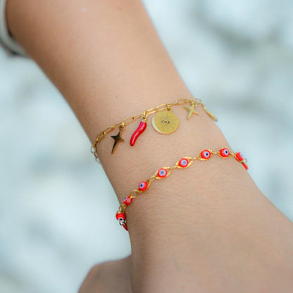 A close-up of a person's wrist wearing two delicate gold-tone bracelets. The top bracelet features small charms including a red chili pepper, a gold disc with an eye motif, and star shapes.