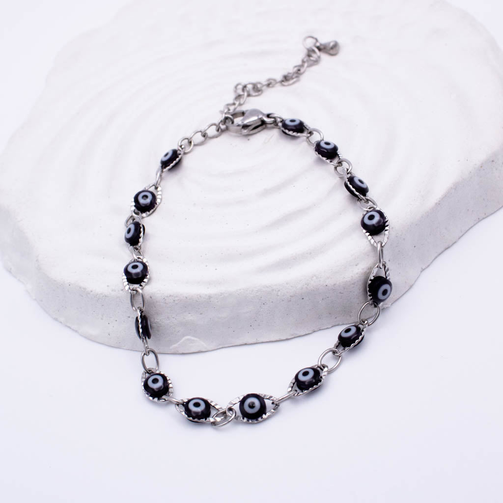 A silver-toned chain bracelet with multiple evil eye beads, displayed on a textured white stone riser against a clean white background.