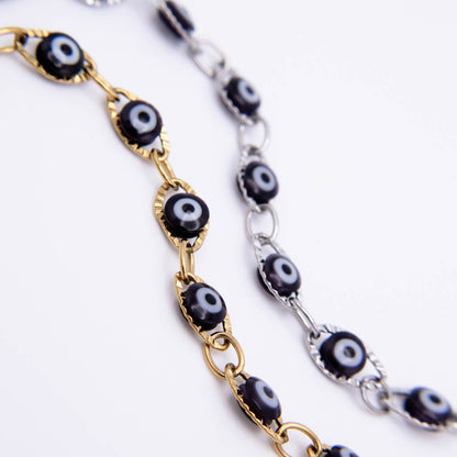 Two evil eye link chains shown on a white background, one in gold-tone metal and one in silver-tone metal, both featuring black, white, and gray circular eye motifs.