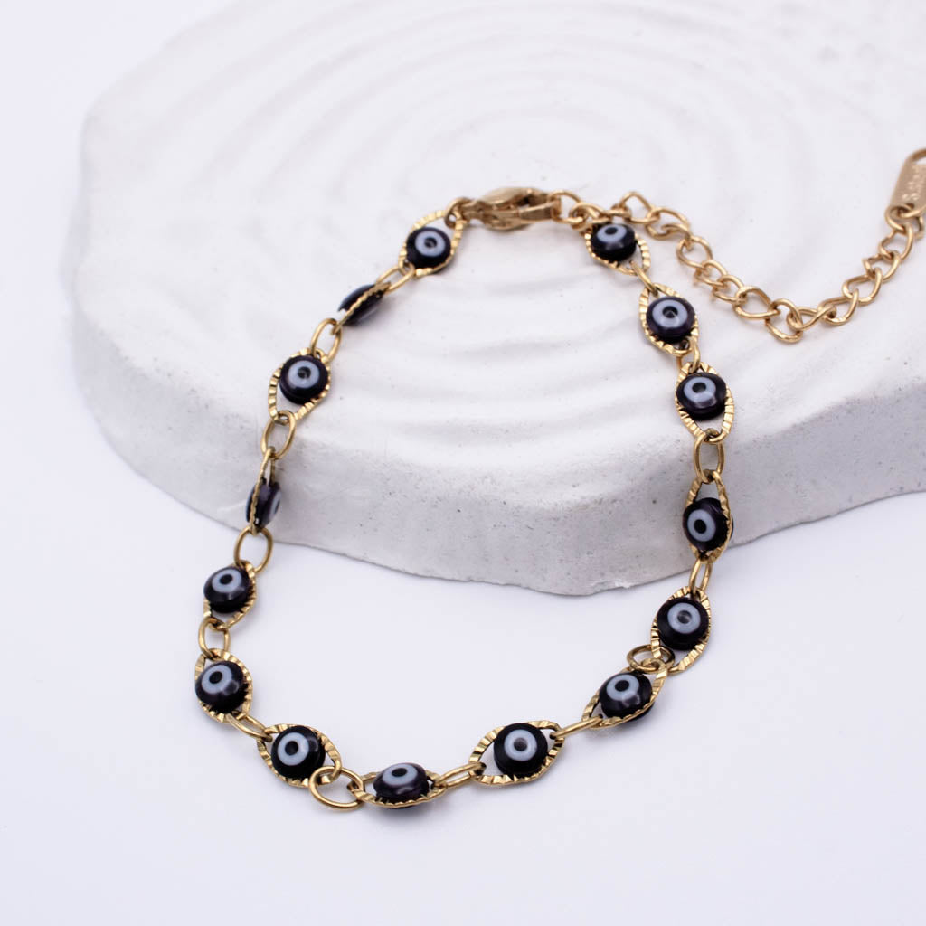 Gold chain bracelet with evil eye beads, displayed on a white textured stone prop against a white background.