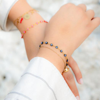 A close-up of a person's wrist wearing multiple delicate bracelets, including a gold chain with blue and white evil eye beads and a small gold charm, alongside other thin bracelets, set against a soft white background.