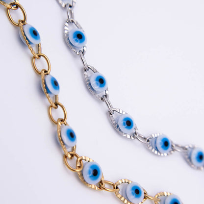 Two close-up views of a chain necklace featuring blue and white evil eye glass beads. One chain is gold-toned, and the other is silver-toned, displayed against a clean white background.