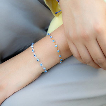 A close-up of a person's wrist wearing two delicate bracelets, one silver and one gold, both adorned with small blue and white evil eye beads. The person is wearing a light yellow top and gray pants.