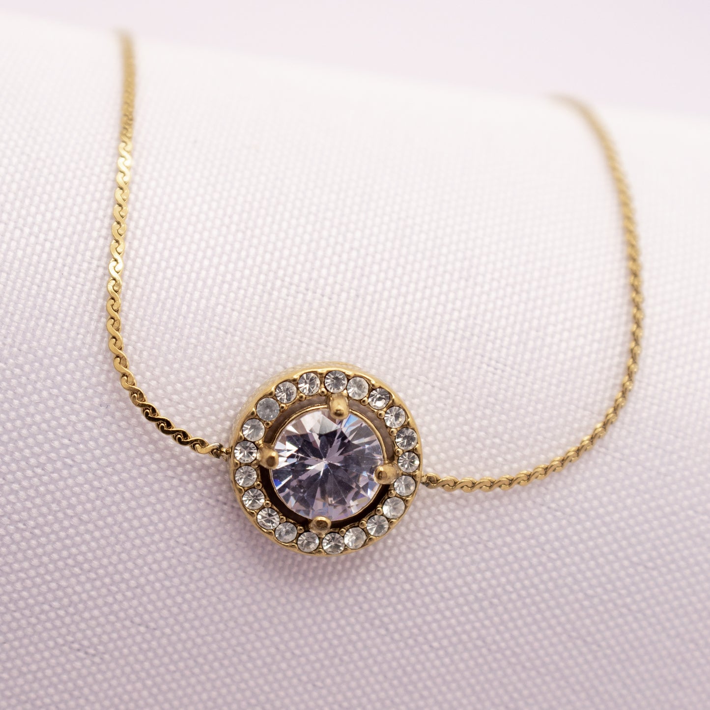 Gold necklace with a circular pendant featuring a large crystal on a light pink background