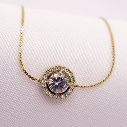 Gold necklace with a circular pendant featuring a large crystal on a light pink background