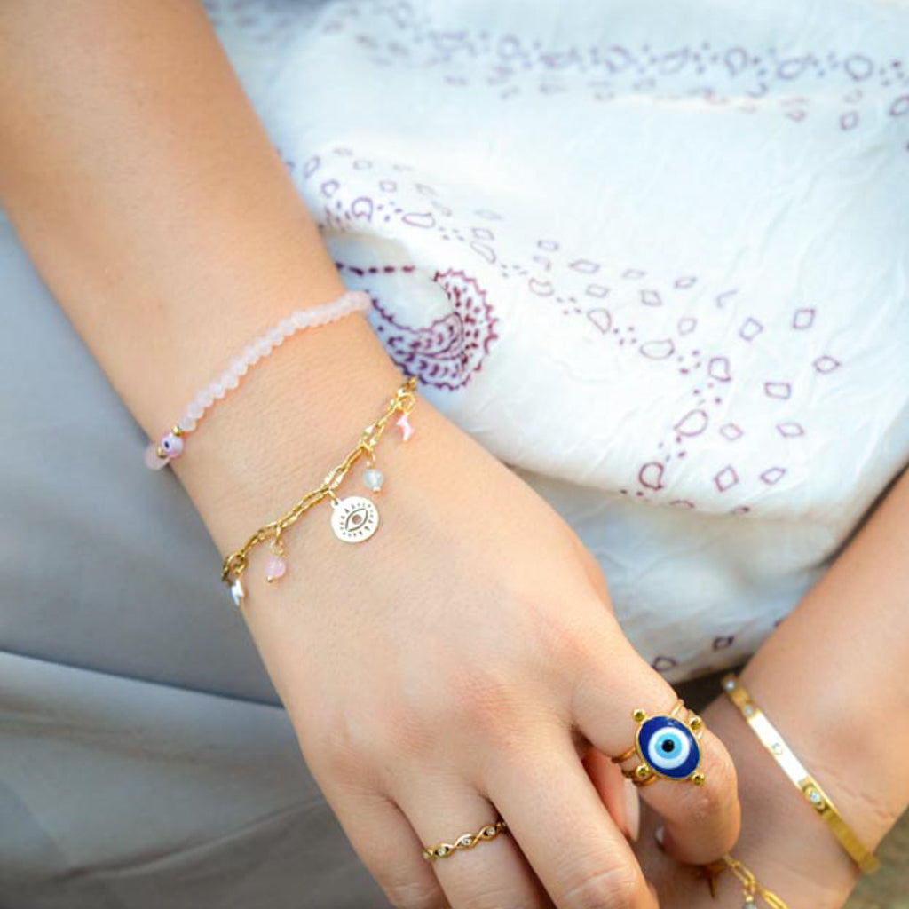 A close-up of a person's hand wearing multiple bracelets and rings, including a gold chain bracelet with a circular charm, a pink beaded bracelet, and a blue-and-white evil eye ring, against a white embroidered top.