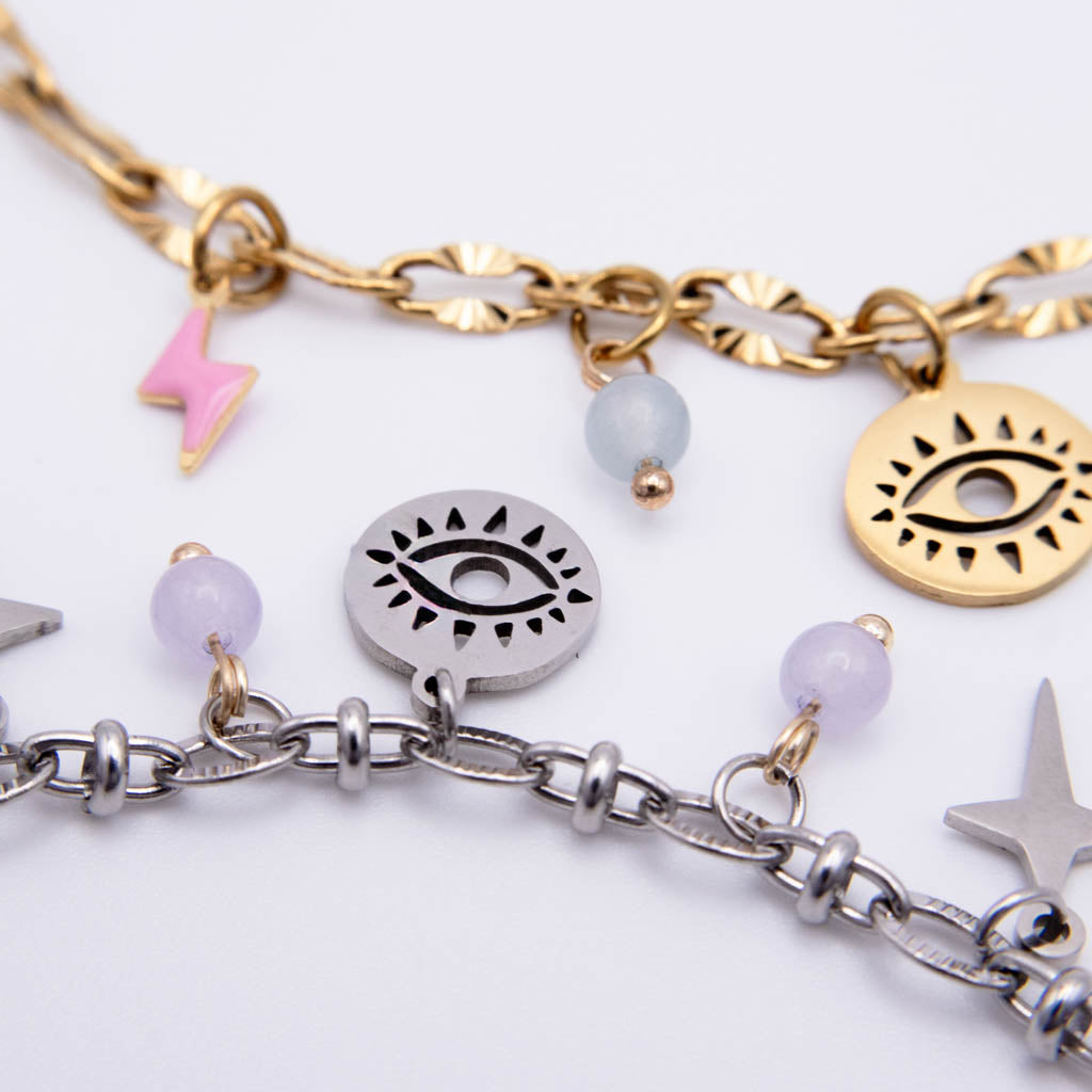 A close-up of a gold and silver chain bracelet with eye, star, and lightning charms, accented by pastel beads on a white background.