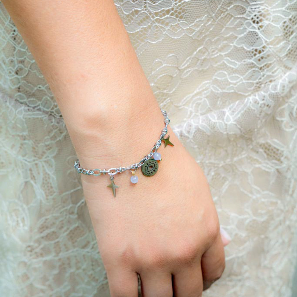 A delicate silver chain bracelet with small charms, including a star, a round medallion, and a cross, worn on a person's wrist against a lace dress background.