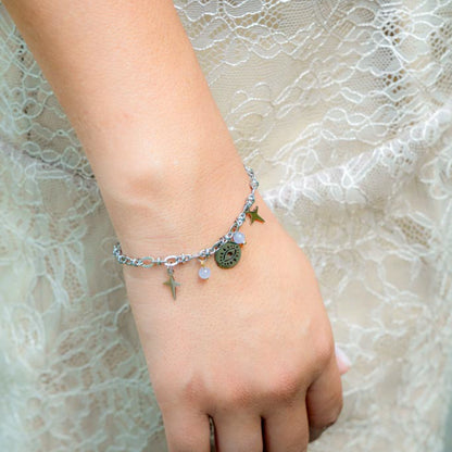 A delicate silver chain bracelet with small charms, including a star, a round medallion, and a cross, worn on a person's wrist against a lace dress background.