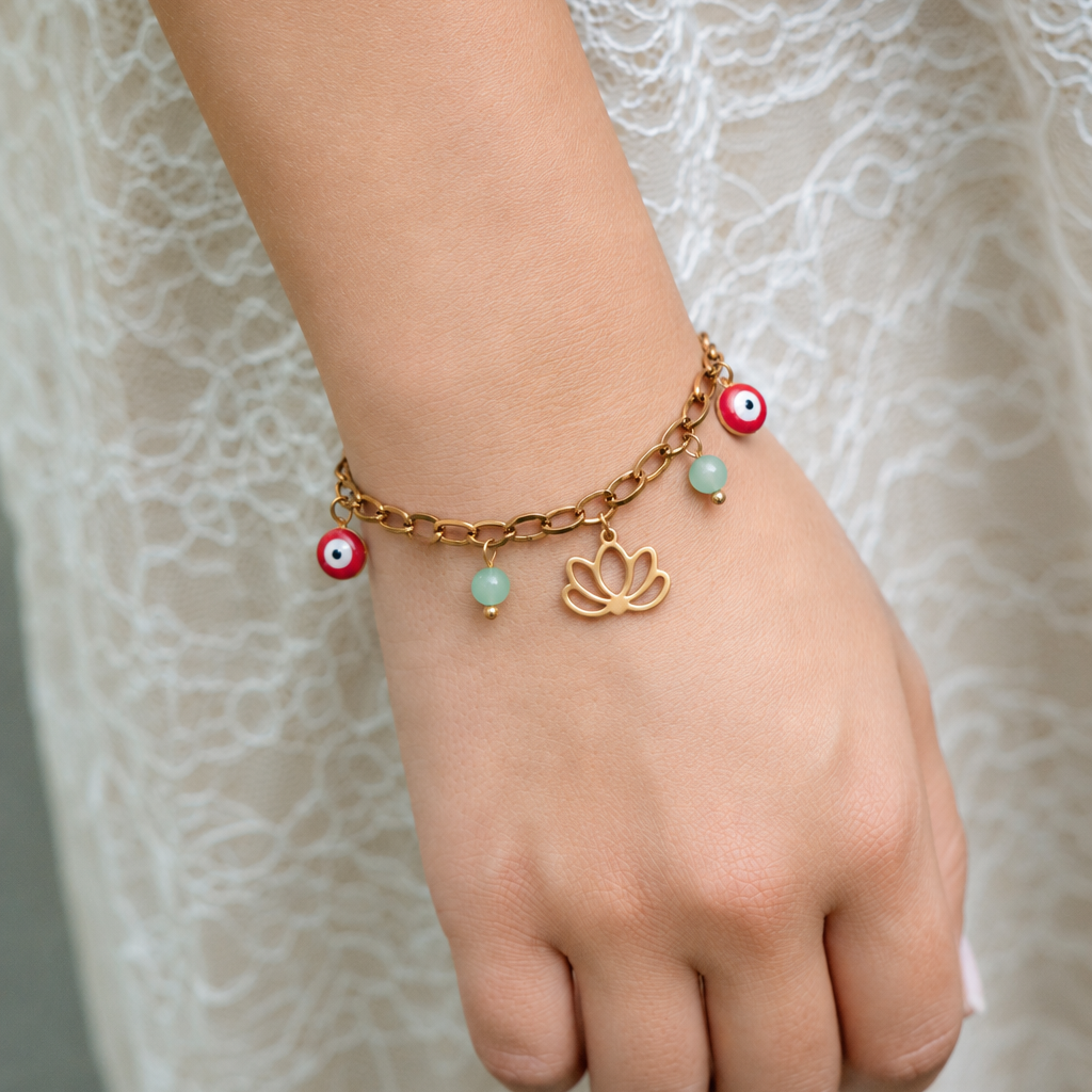 A gold chain bracelet with evil eye charms, mint green beads, and a lotus flower charm, shown worn on a person's wrist against a white lace dress background.