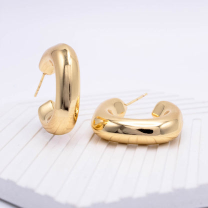 A pair of polished gold huggie hoop earrings with a chunky, rounded modern design, displayed on a white textured surface.