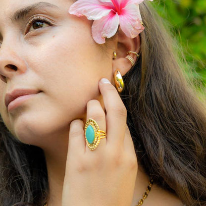 A close-up lifestyle shot of a woman wearing a gold ring with a large oval turquoise cabochon, along with other gold earrings and a flower in her hair.