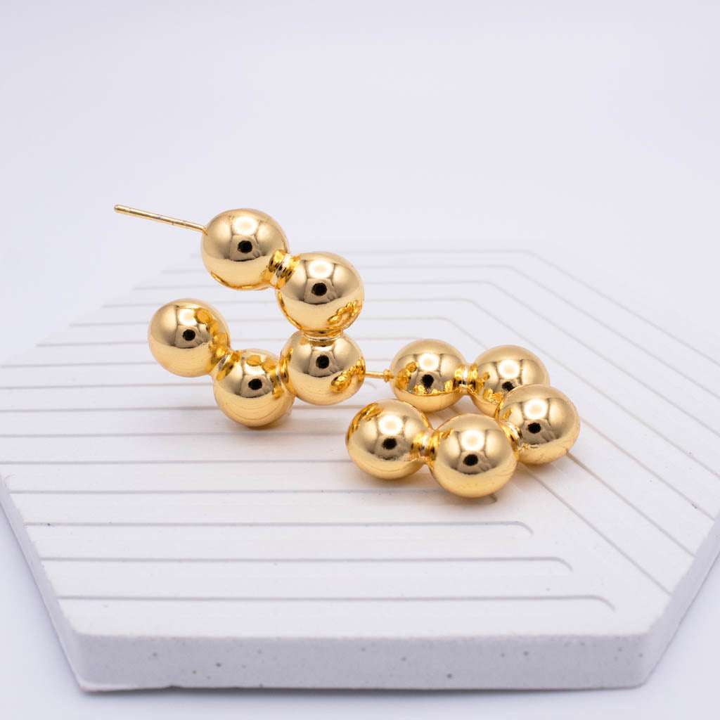 Gold beaded drop earrings with multiple polished spherical beads on each earring, displayed on a white textured hexagonal surface against a light gray background.