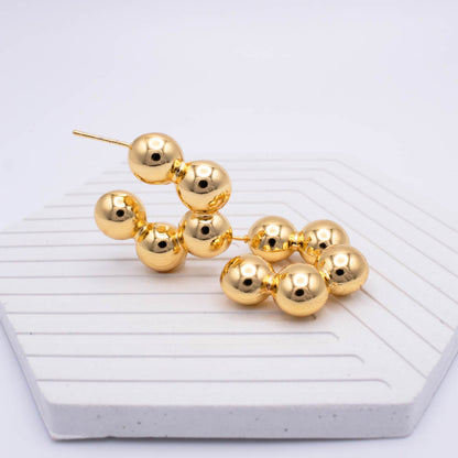 Gold beaded drop earrings with multiple polished spherical beads on each earring, displayed on a white textured hexagonal surface against a light gray background.