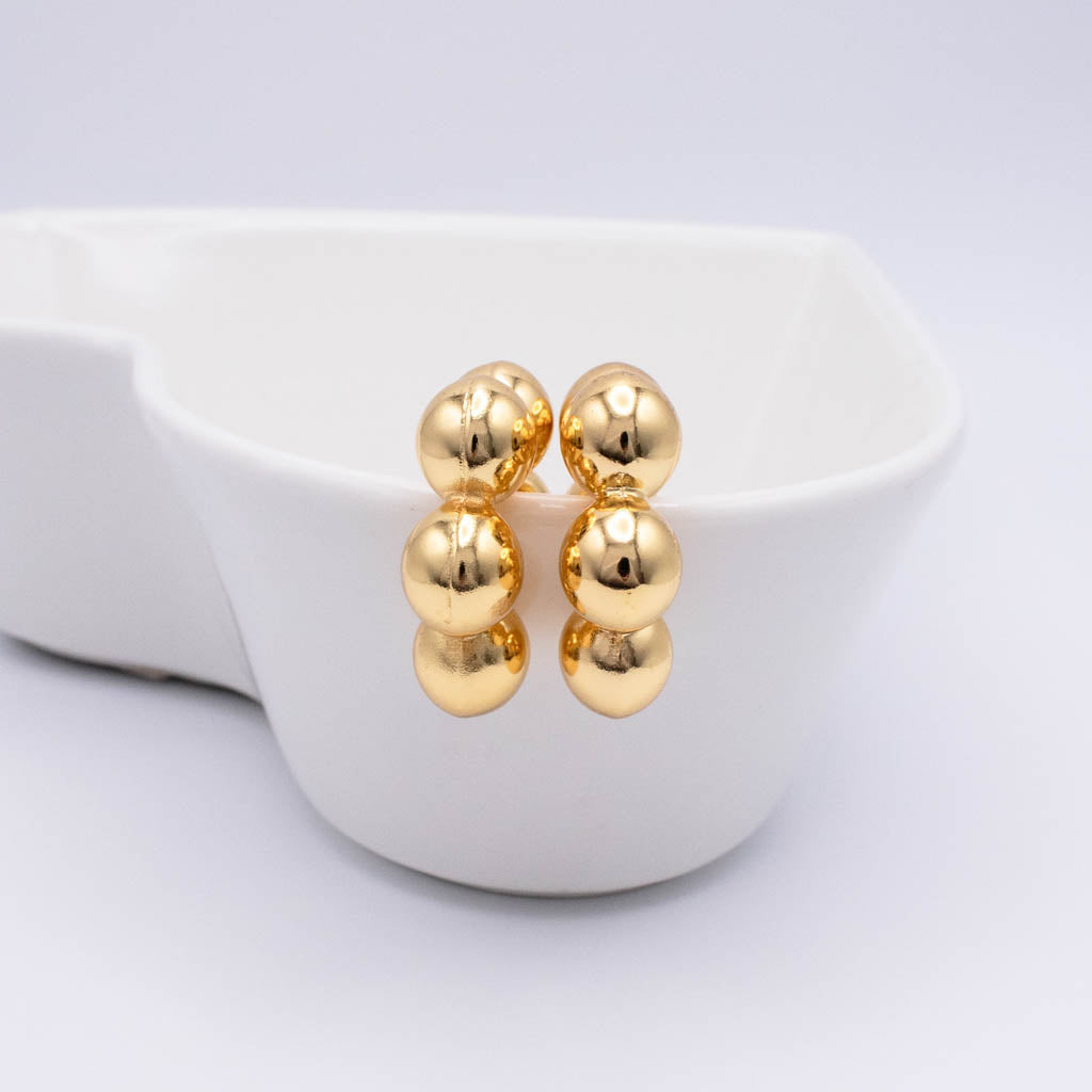 A pair of gold stud earrings with three smooth, rounded bead-like spheres stacked vertically, displayed on the rim of a white ceramic dish against a light gray background.