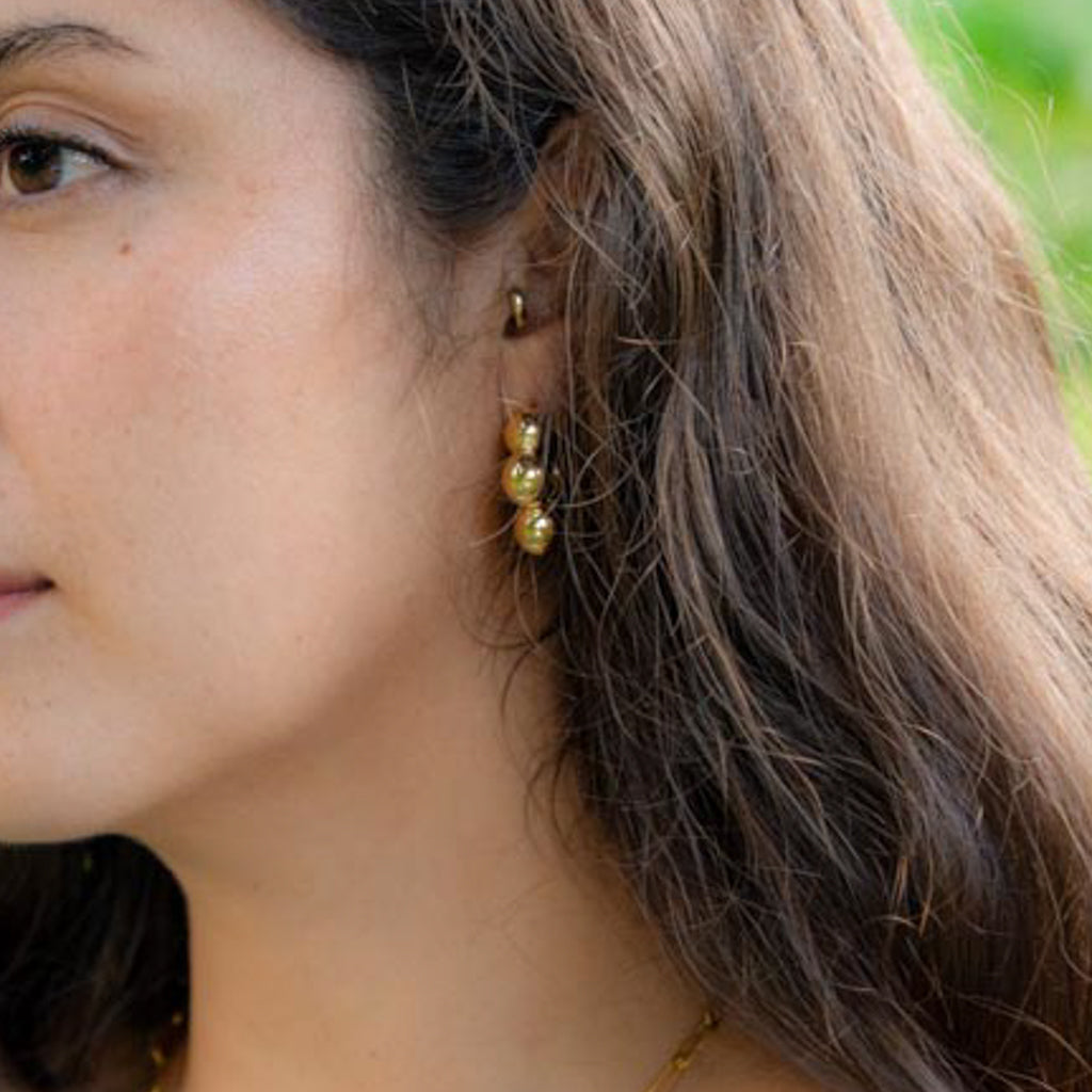 A close-up side profile of a woman wearing a small gold hoop earring with three dangling gold beads, set against her dark brown hair and a soft green blurred background.