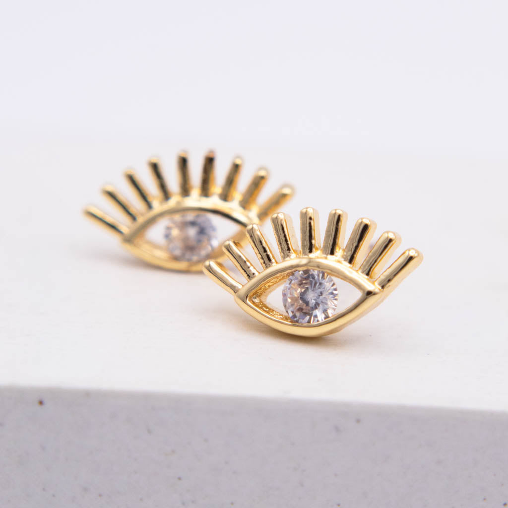 A pair of gold eye-shaped stud earrings with clear gemstones in the center, displayed on a clean white surface.
