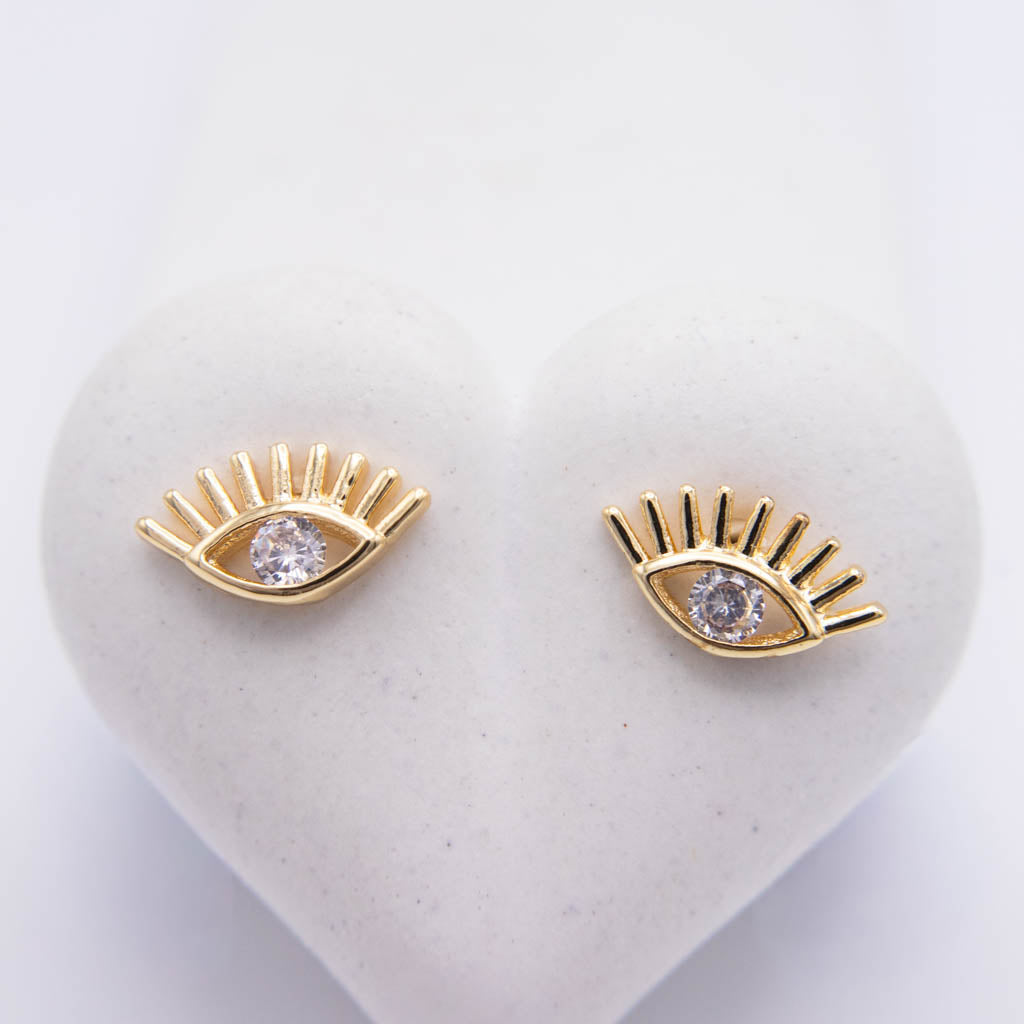 A pair of gold eyelash stud earrings with clear round gemstones, displayed on a white heart-shaped jewelry stand against a white background.