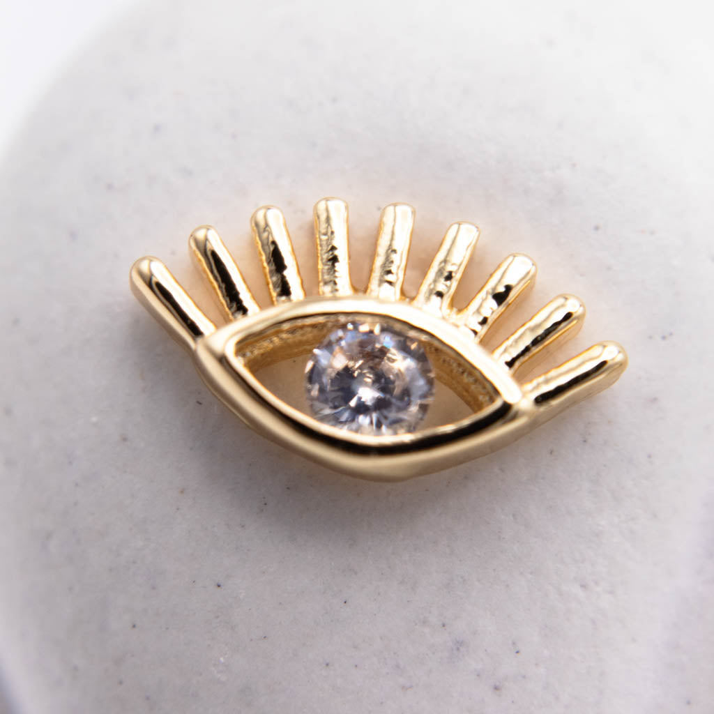 A gold-plated evil eye charm with a clear faceted gemstone in the center, resting on a light gray textured surface.