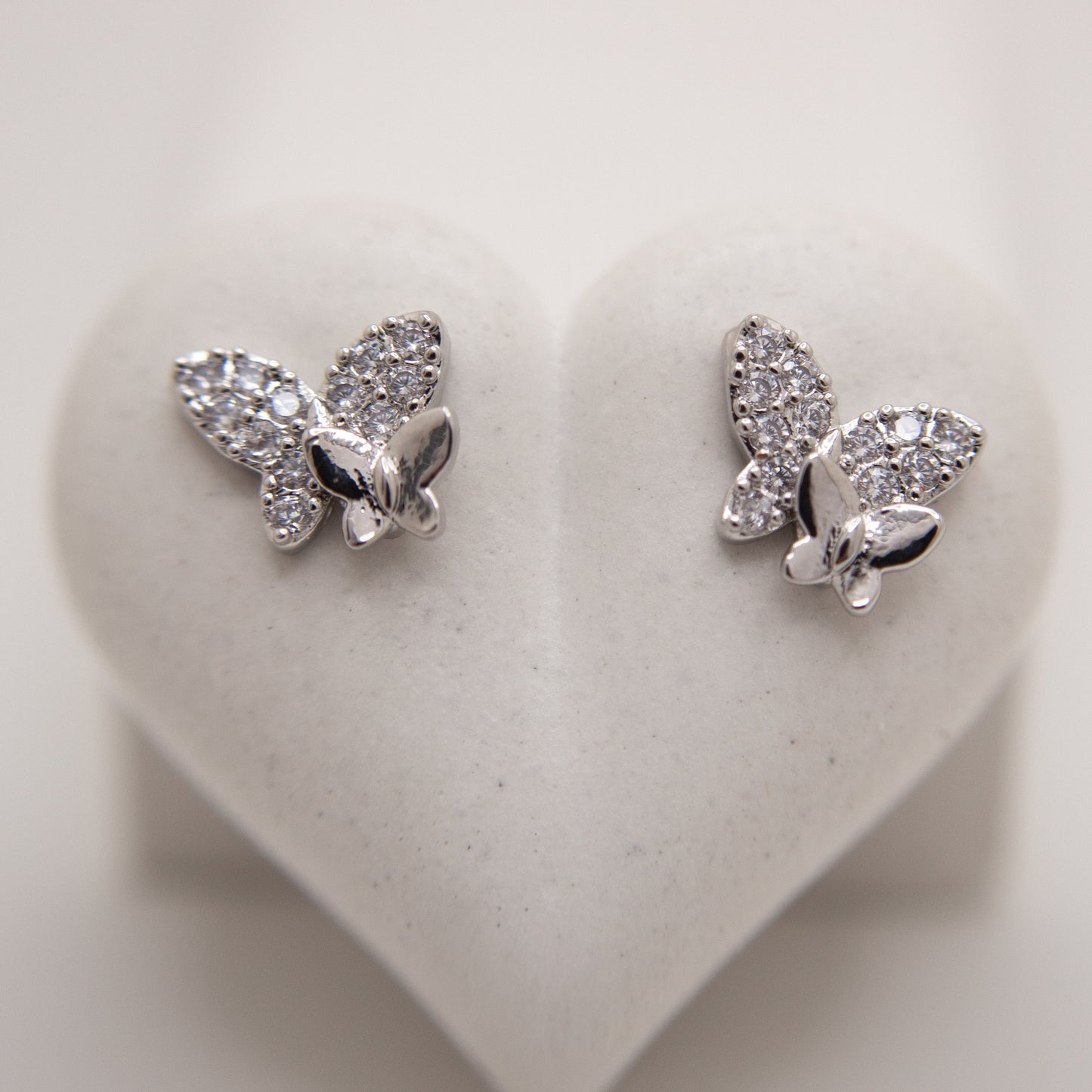 Butterfly-shaped silver earrings with clear stones on a heart-shaped white background