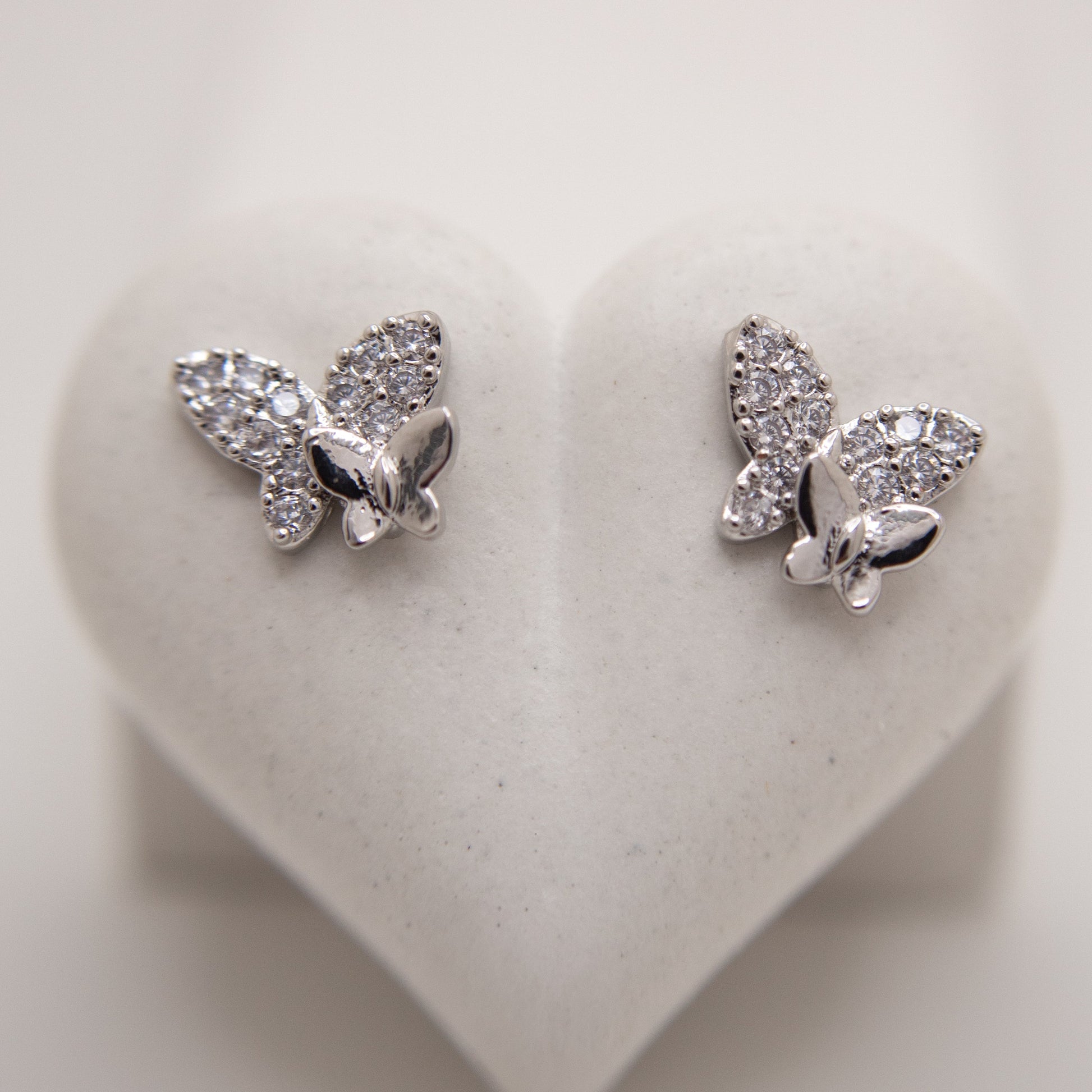 Butterfly-shaped silver earrings with clear stones on a heart-shaped white background