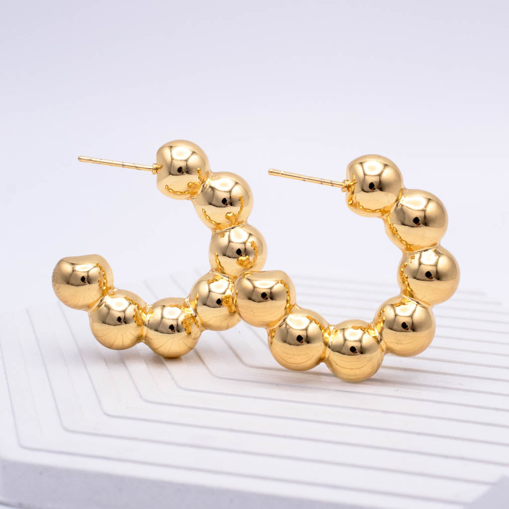 Gold hoop earrings made of connected glossy round beads, displayed on a white textured surface against a light gray background.