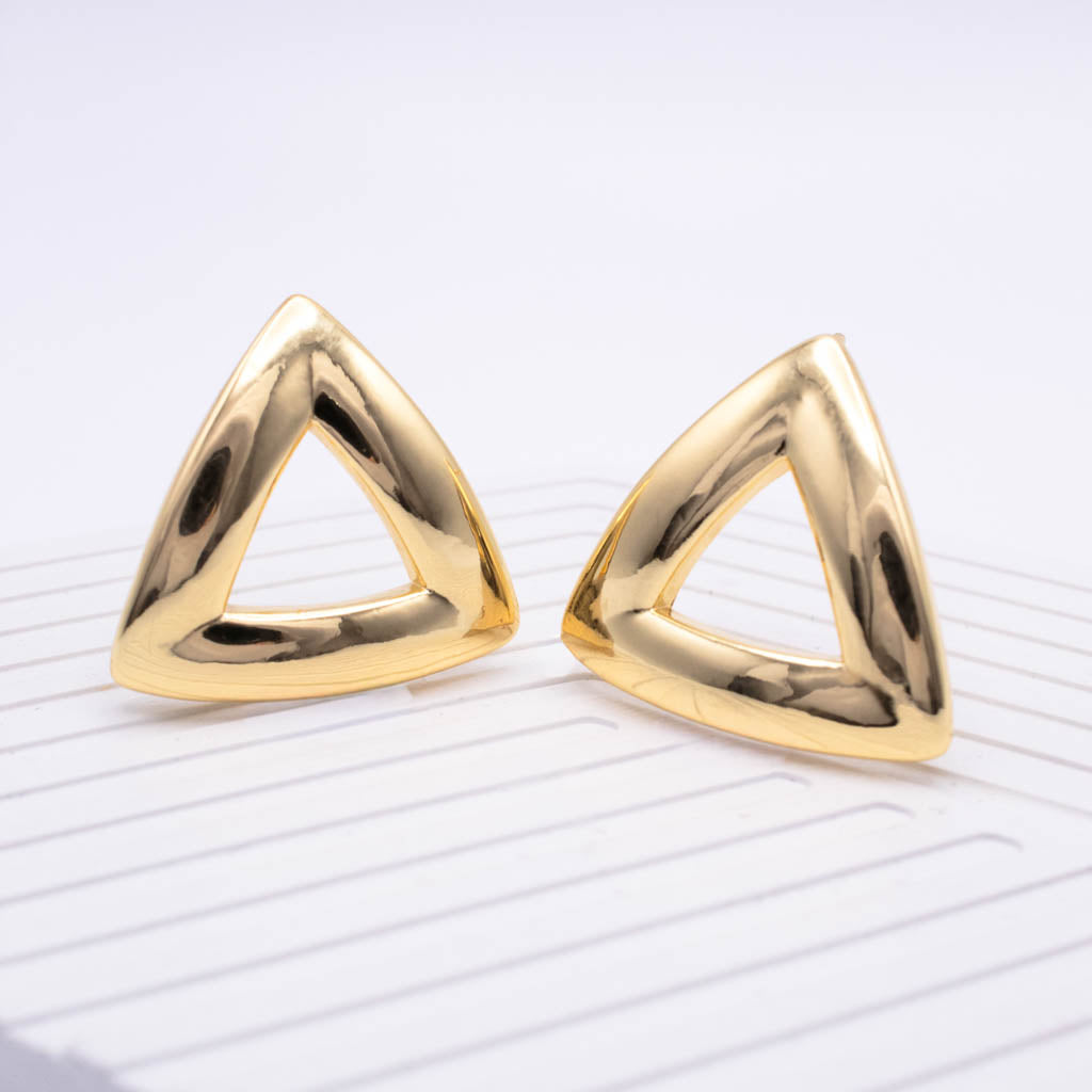 A pair of polished gold triangular stud earrings with a thick, open triangular frame design, displayed on a white textured surface.