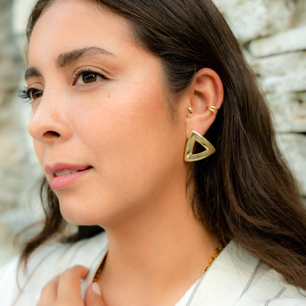 A close-up of a woman wearing a gold triangular hoop earring, with small gold ear cuffs and a gold necklace, set against a blurred stone wall background.