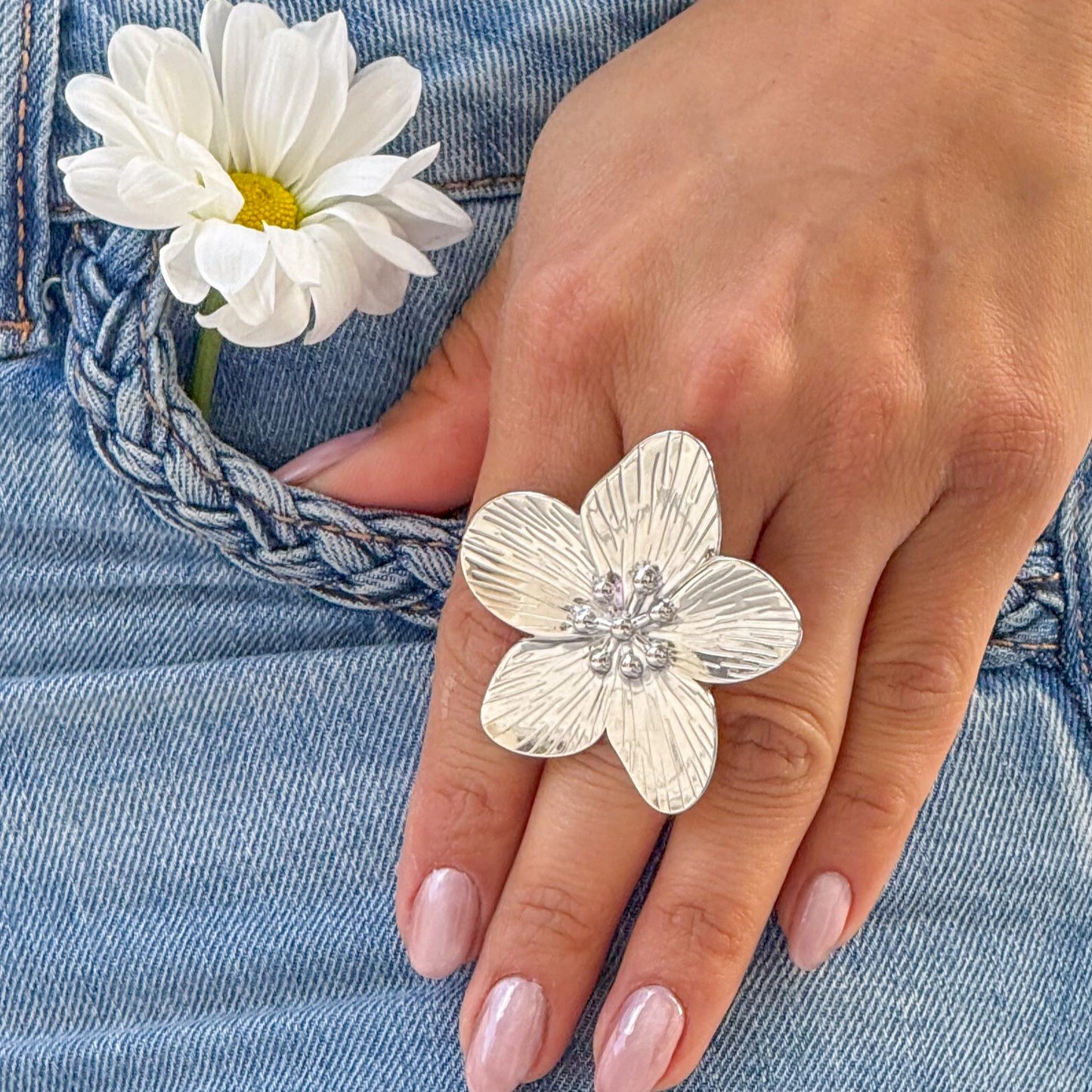 Hand wearing a silver floral ring with a white flower in the background