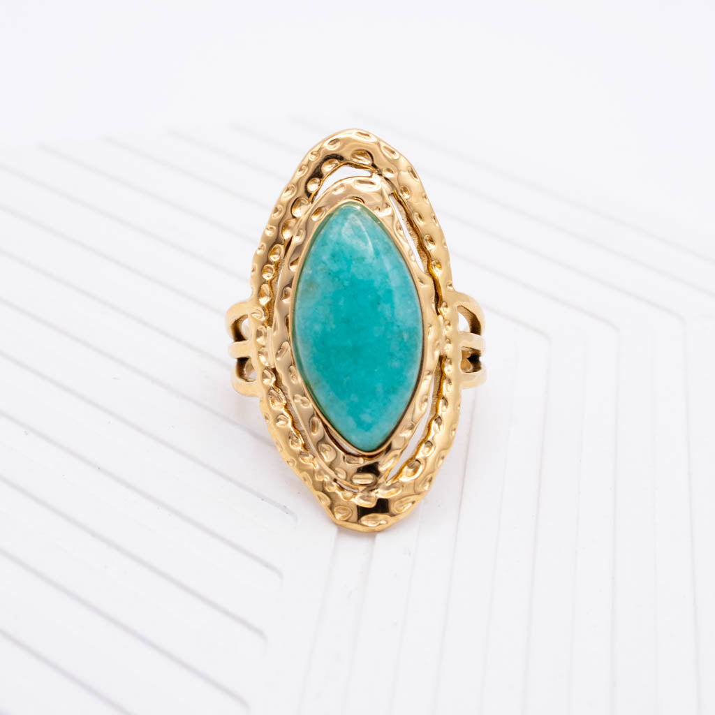 A gold ring featuring a large marquise-shaped turquoise stone in an ornate textured setting, photographed on a clean white background with subtle diagonal lines.