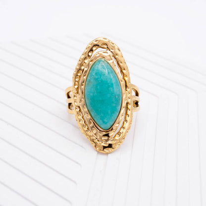A gold ring featuring a large marquise-shaped turquoise stone in an ornate textured setting, photographed on a clean white background with subtle diagonal lines.