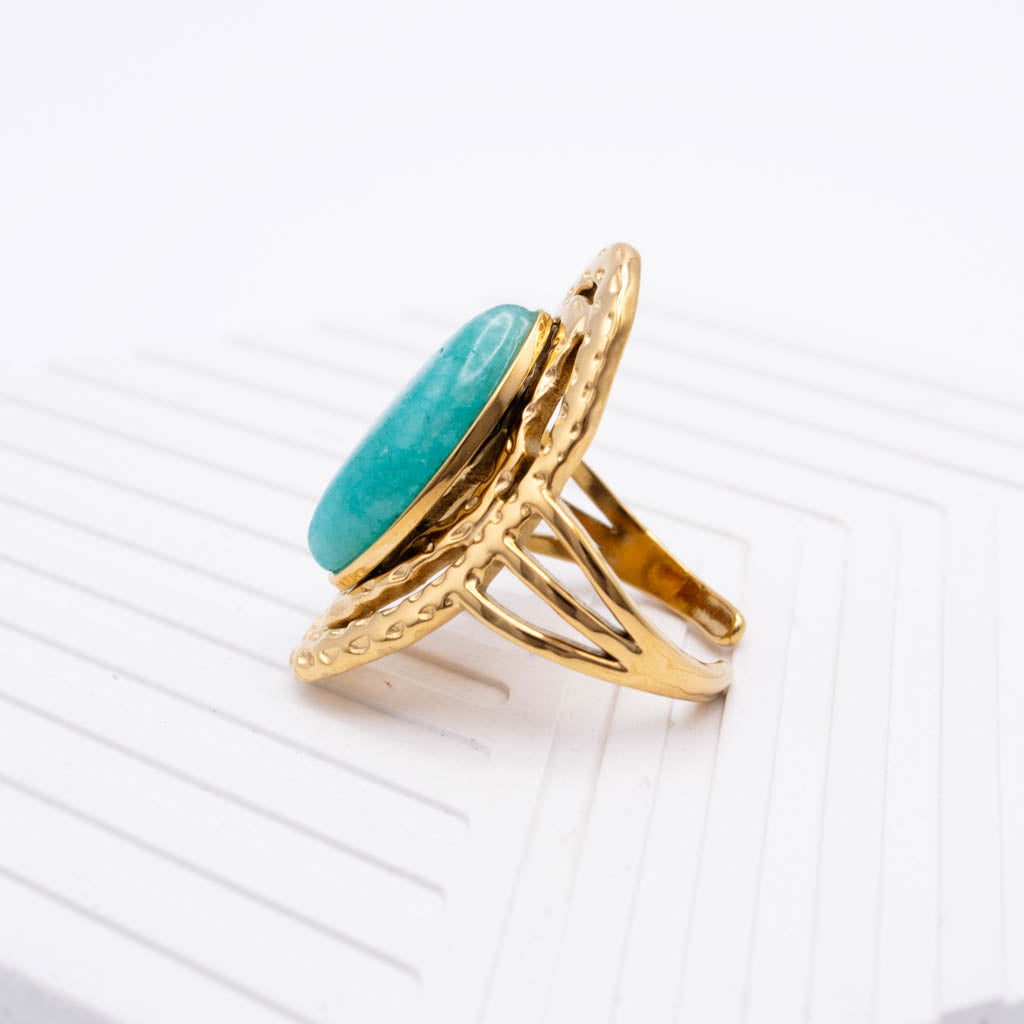 A gold ring with a large oval turquoise-colored stone, displayed on a white textured surface.