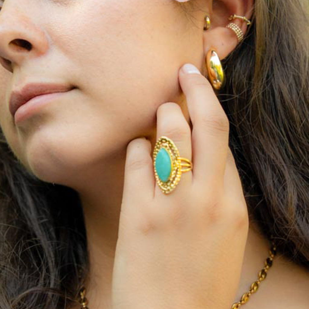 A close-up lifestyle shot of a woman wearing a gold ring with a large oval turquoise stone, along with matching gold earrings and a necklace.