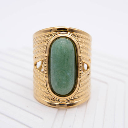 A wide gold ring with a textured, basketweave-like band and a large oval green stone in the center, shown on a white textured background.