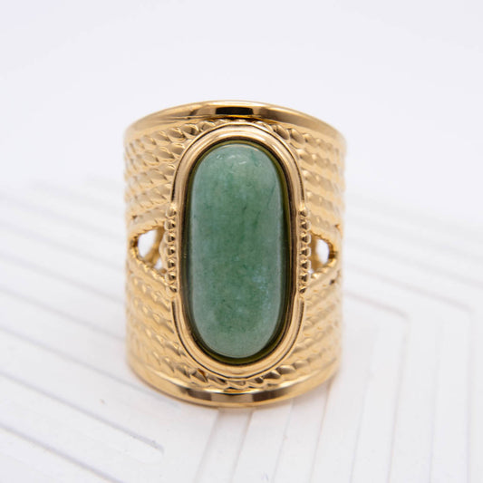 A wide gold ring with a textured, basketweave-like band and a large oval green stone in the center, shown on a white textured background.