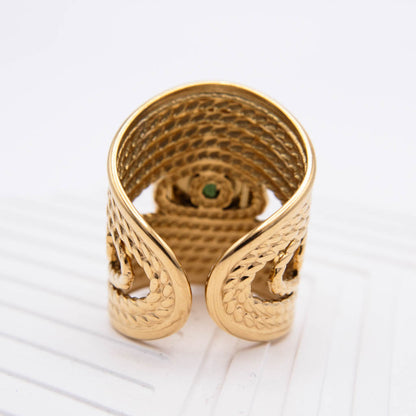 A gold-toned wide band ring with a textured, woven-like outer surface and a small green stone set in the center, photographed on a clean white background.