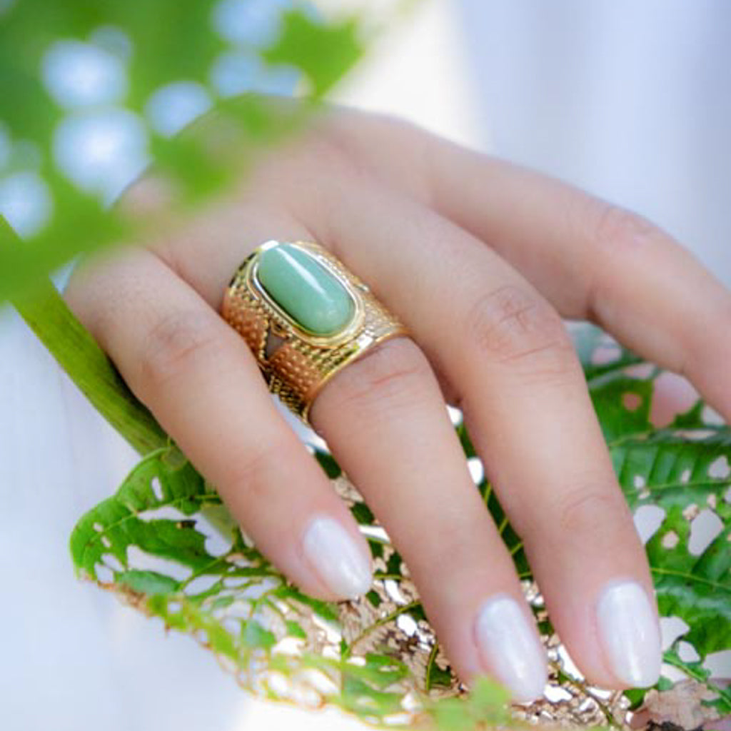 A close-up of a hand wearing a gold ring with a smooth, light green oval stone, resting on green leaves.