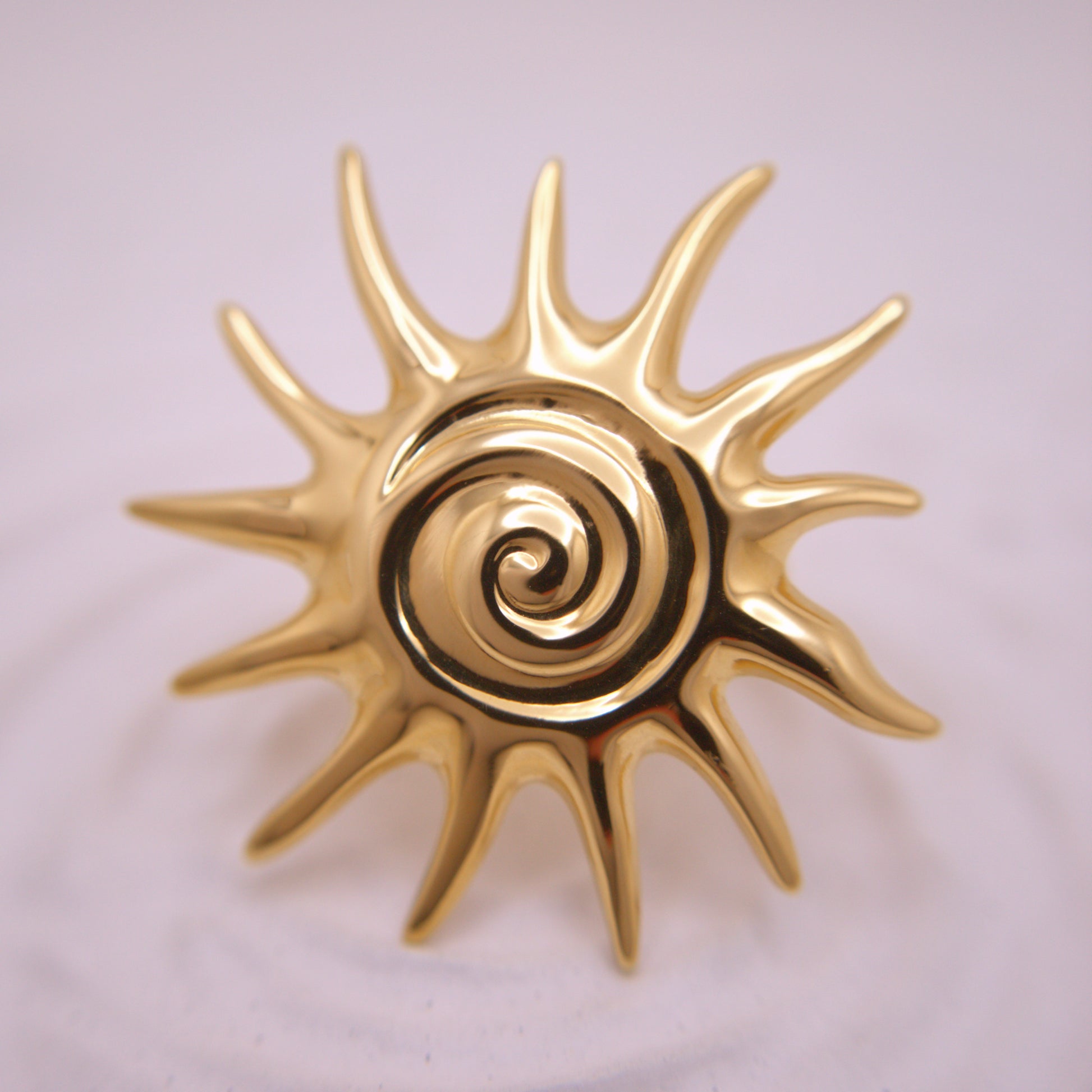 Gold sun-shaped decorative item with a central spiral design on a light pink background
