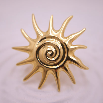 Gold sun-shaped decorative item with a central spiral design on a light pink background