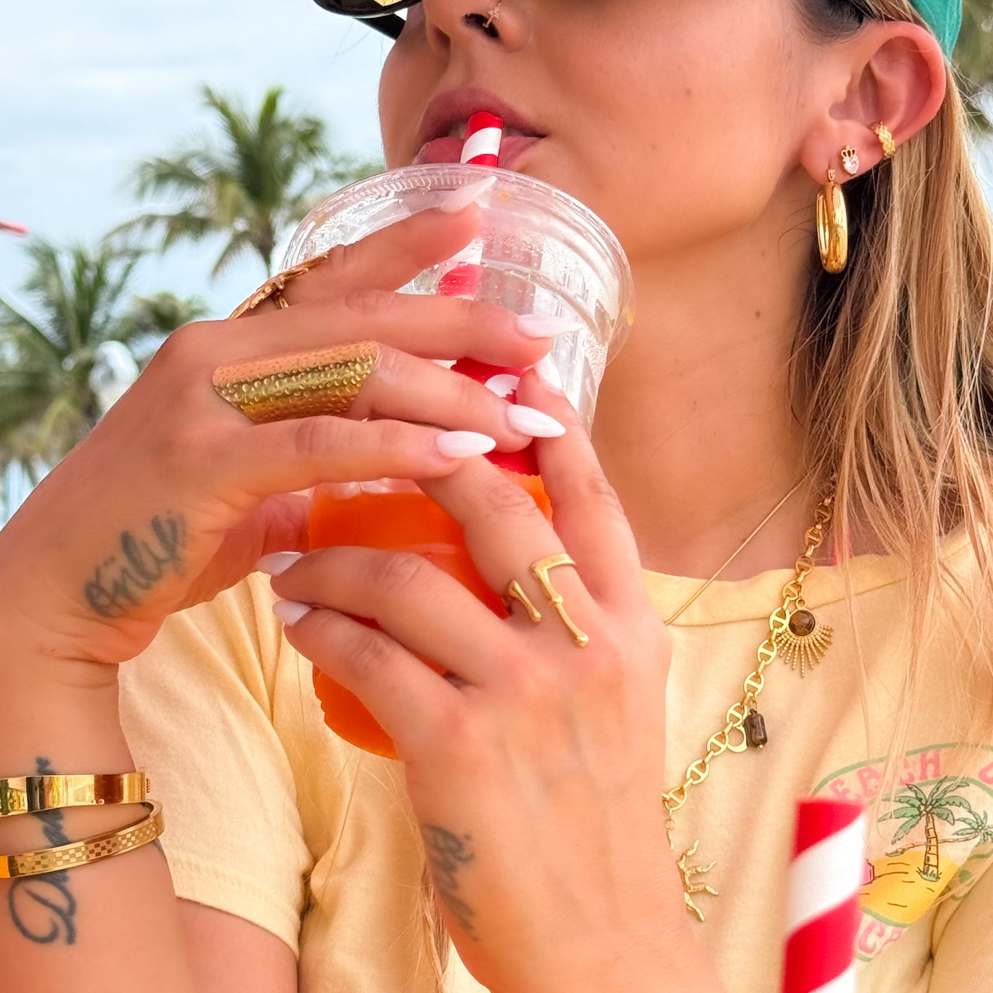 Woman drinking from a red cup with a straw, wearing gold jewelry and a yellow shirt.