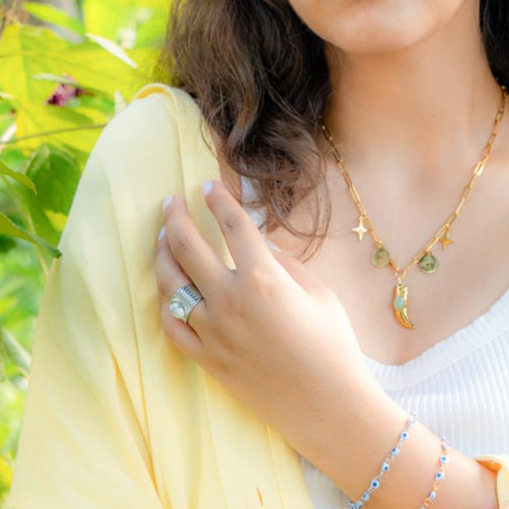 A close-up lifestyle shot of a woman wearing a gold celestial necklace with a crescent moon and star pendants, a silver ring, and a blue beaded bracelet, set against a background of green foliage and a pale yellow garment.