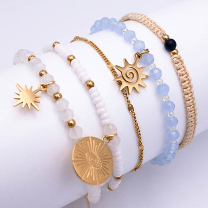 A set of five bohemian-style beaded and chain bracelets displayed on a white cylindrical jewelry stand. The bracelets feature white and light blue beads, gold-toned metal chains and charms (including sun and moon designs), a white stone bead bracelet with a gold disc charm, a white beaded bracelet with a gold star charm, and a tan cord bracelet with a black bead.