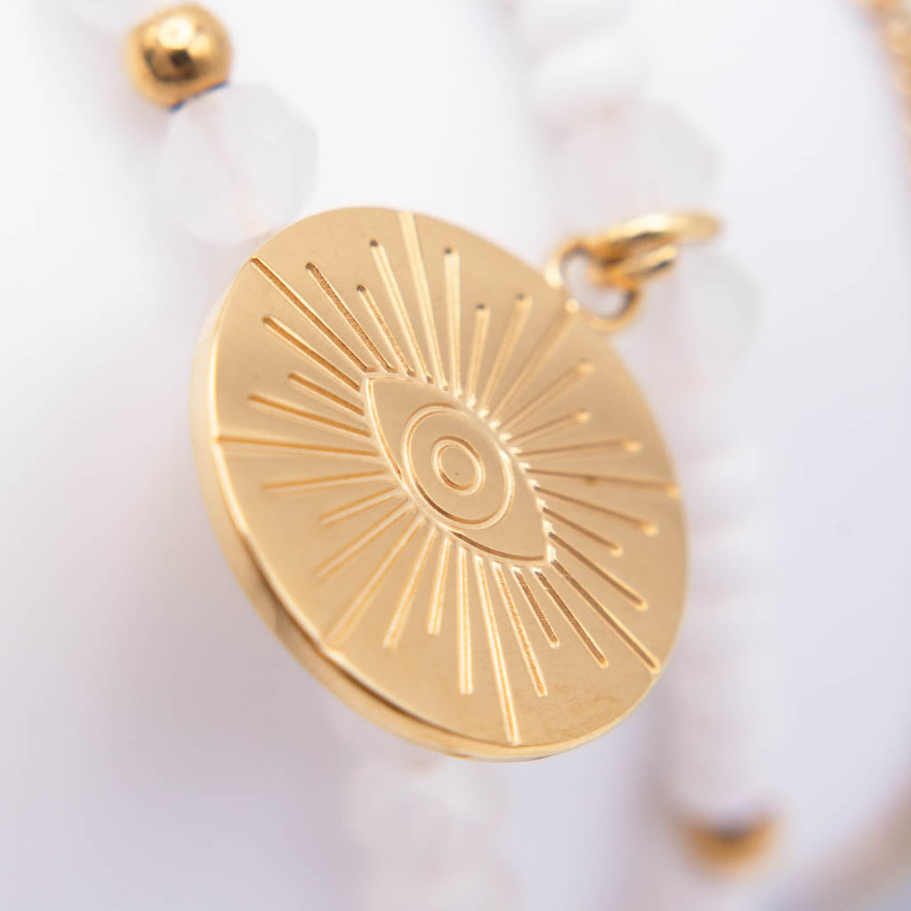 A close-up of a gold oval pendant featuring an engraved sunburst and eye design, hanging from a white beaded necklace with white beads and gold accents.