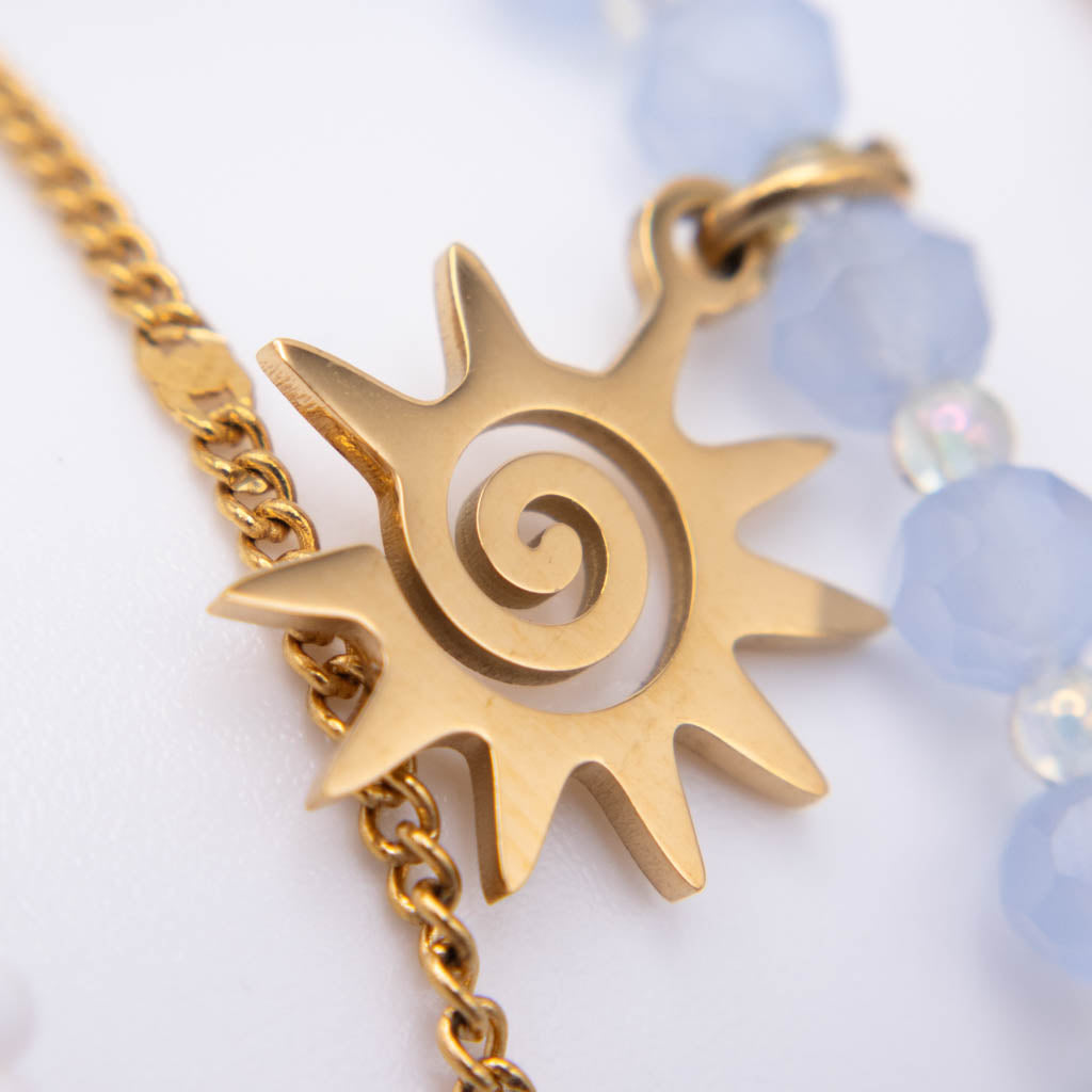 A gold sun pendant charm with a spiral cutout, hanging from a gold chain next to a strand of light blue faceted beads on a white background.