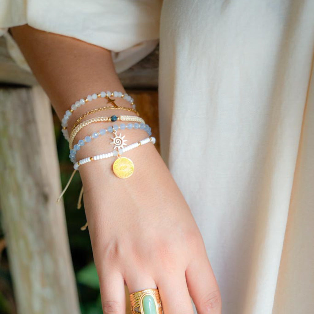 A close-up of a person's wrist wearing multiple delicate beaded bracelets, including light blue, white, and gold-toned strands with a small yellow sun charm, paired with a gold ring.