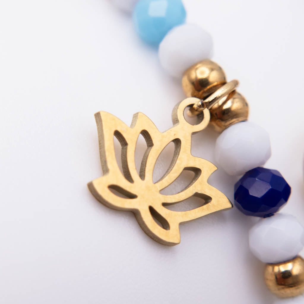 Close-up of a beaded jewelry strand with a gold lotus-shaped charm, white and light blue faceted beads, and small gold bell charms on a white background.