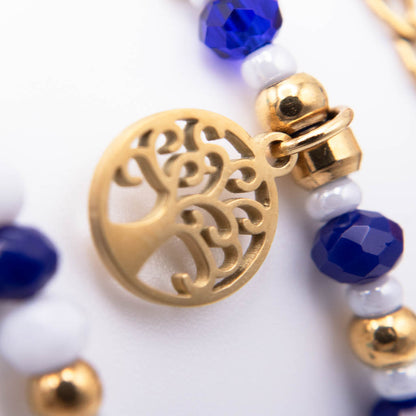 Close-up of a beaded bracelet featuring a gold tree of life pendant, faceted blue beads, and white beads on a white background.