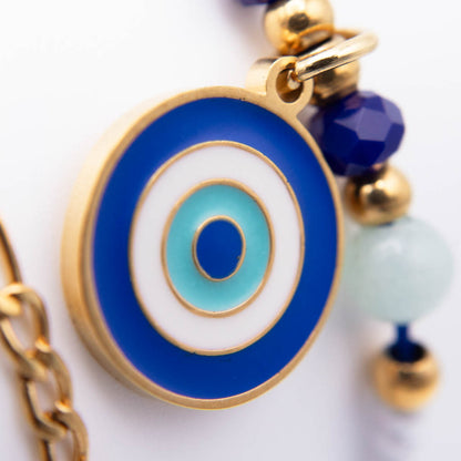 A close-up of a gold-tone evil eye charm with concentric blue, white, and light blue enamel rings, attached to a gold chain with blue and pale green beads.