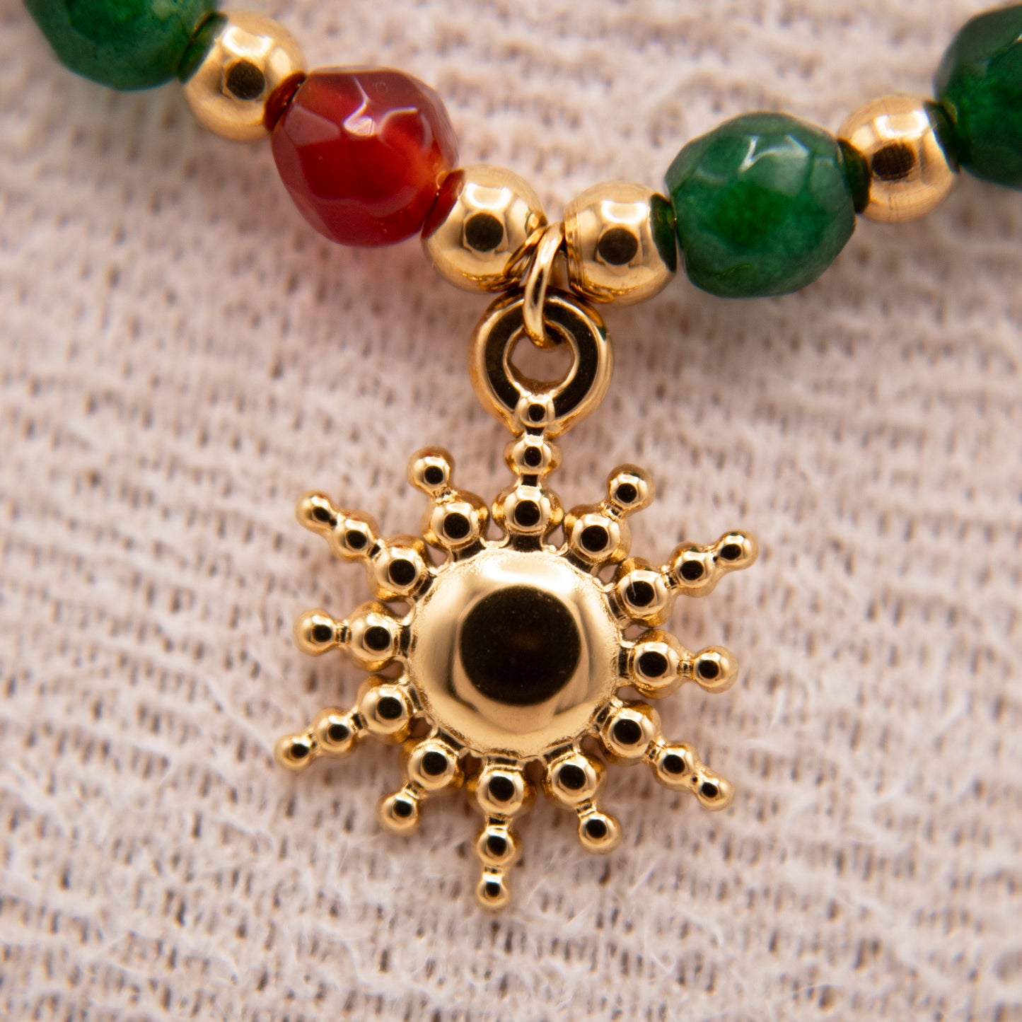 Gold sun-shaped pendant on a beaded bracelet with green and red beads on a textured beige background