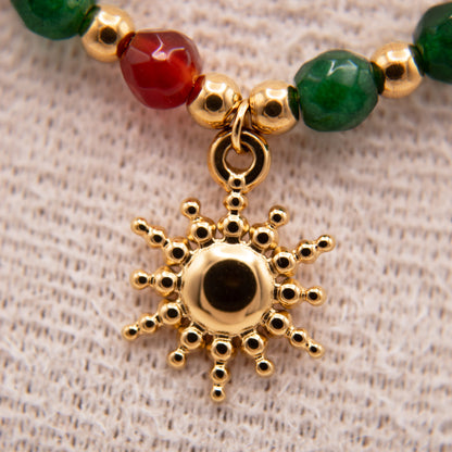 Gold sun-shaped pendant on a beaded bracelet with green and red beads on a textured beige background